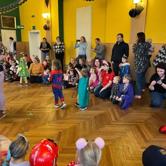 Eine Gruppe von Kindern und Erwachsenen in Kostümen befindet sich in einem Raum, einige sitzen auf dem Boden, während andere stehen und zusehen. Der Raum hat gelbe Wände und einen Holzboden.