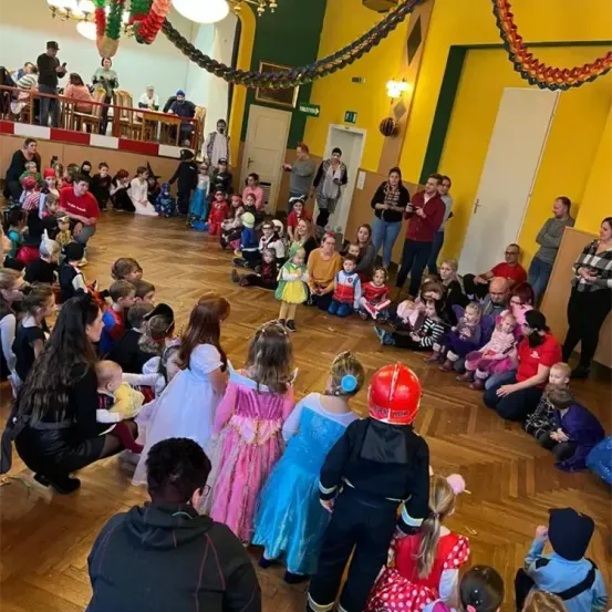 Eine Gruppe von Kindern in Kostümen versammelt sich in einem Saal mit Holzböden, Erwachsene stehen und sitzen um sie herum. Einige sind als Superhelden und Feuerwehrleute verkleidet.