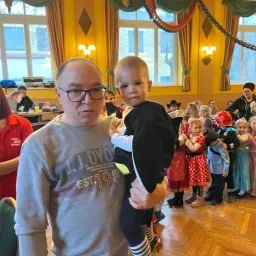 Ein Mann hält ein Baby in einem Raum mit Kindern in Kostümen. Sie stehen in einer Reihe und schauen das Baby an. Der Raum hat gelbe Wände und Holzböden.