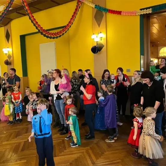 Eine Gruppe von Menschen versammelt sich in einem Raum. Einige Kinder tragen Kostüme. Der Raum ist mit buntem Papier dekoriert. Eine Frau in einem roten Shirt steht vor ihnen.