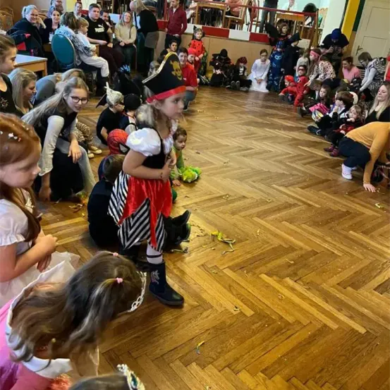 Ein junges Mädchen in einem Piratenkostüm steht inmitten eines Raumes mit Kindern in verschiedenen Kostümen. Der Raum hat einen Holzboden und eine Gruppe von Menschen, einige sitzend und andere stehend, beobachten.