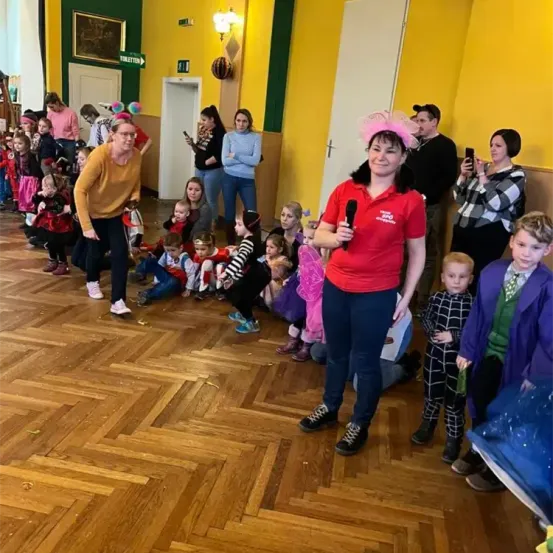 Eine Gruppe von Menschen ist in einem Raum, eine Frau in Rot hält ein Mikrofon, und Kinder sind in verschiedenen Kostümen, einige sitzen auf dem Boden und andere stehen.