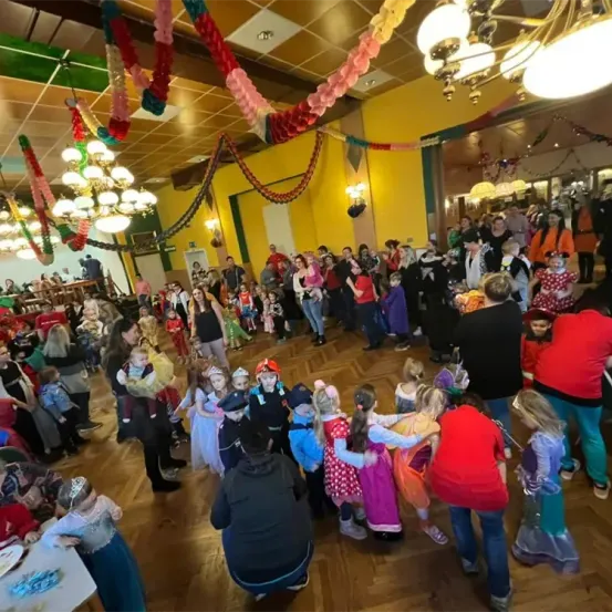 Ein großer Raum mit Holzboden ist mit Menschen gefüllt, darunter Kinder in Kostümen und Erwachsene, einige halten Babys. Dekorationen und Lichter schmücken den Raum.
