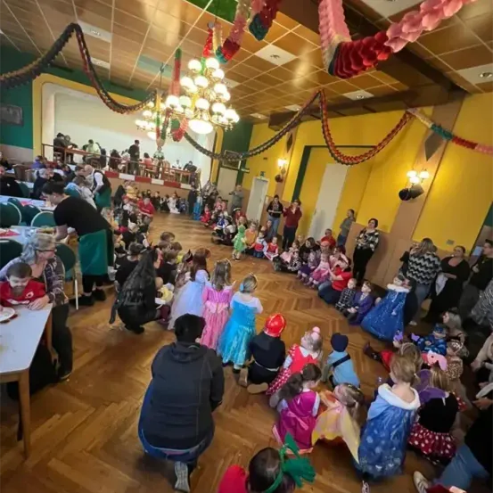 Eine Gruppe von Kindern und Erwachsenen sitzt in einem Kreis in einem dekorierten Raum. Einige Kinder tragen Kostüme. Es gibt eine Kronleuchter und einen Tisch mit Tellern.