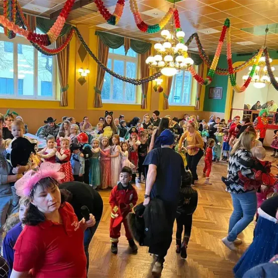 Eine festliche Zusammenkunft von Kindern und Erwachsenen in einem geschmückten Saal, viele in Kostümen gekleidet, mit einer Frau vorne, die ein Mikrofon hält, und einem Mann, der eine Tasche trägt.