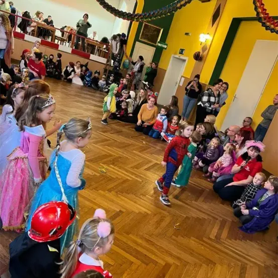 Kinder in Kostümen versammeln sich in einem Saal, einige sitzen auf dem Boden, während andere stehen. Ein Junge in einem Spider-Man-Kostüm läuft auf das Publikum zu. Oben blickt eine Galerie auf die Szenerie herab.