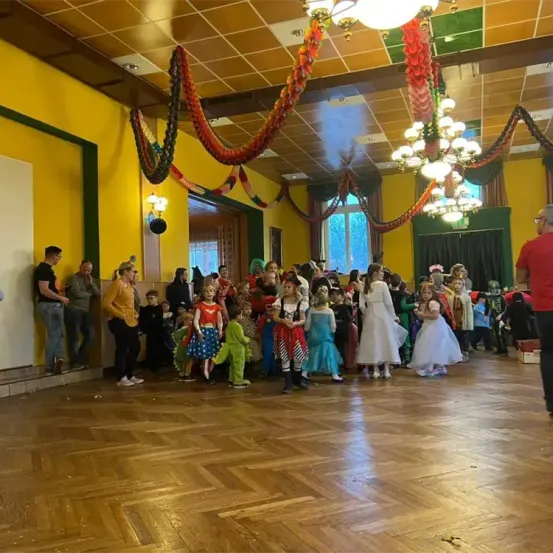 In einem Saal mit Holzböden versammeln sich Kinder in Kostümen mit Erwachsenen, beleuchtet von Kronleuchtern, umgeben von farbenfrohen Dekorationen.
