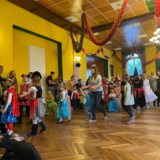 Kinder in verschiedenen Kostümen tanzen in einem für ein Fest dekorierten Raum mit gelben Wänden und festlichen Dekorationen. Einige tragen Superhelden-Capes, andere Prinzessinnenkleider.