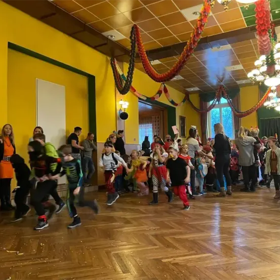 Eine Gruppe von Kindern in Kostümen tanzt auf einem Holzboden, umgeben von Erwachsenen. Der Raum ist mit bunten Girlanden und Lichtern dekoriert.