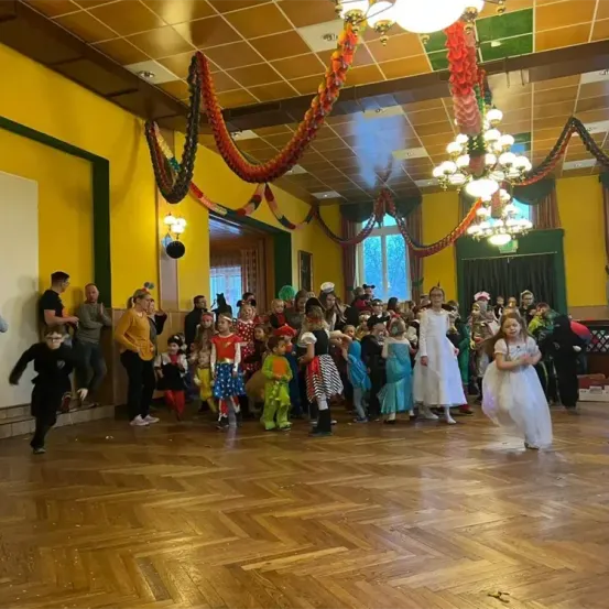 Eine Gruppe von Kindern in verschiedenen Kostümen steht auf einem Holzboden. Sie versammeln sich in einem Raum mit gelben Wänden und einer an der Decke hängenden Kronleuchter. Einige Erwachsene stehen an der Seite und beobachten die Kinder.