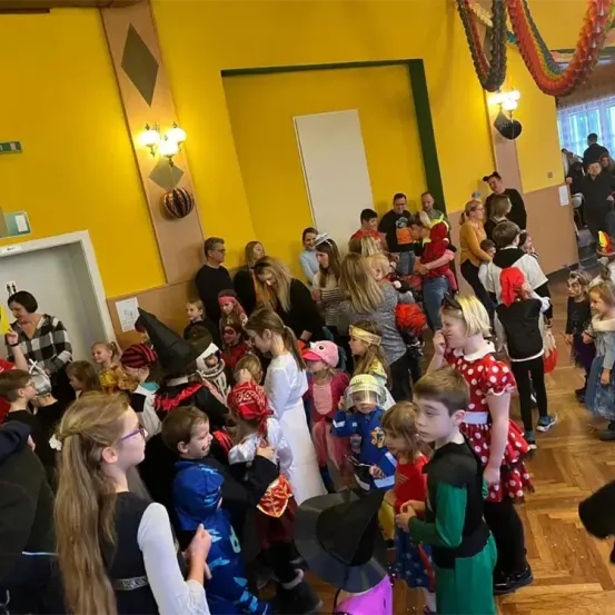 Eine Gruppe von Kindern und Erwachsenen in einem Raum, viele in Kostümen gekleidet. Der Raum hat gelbe Wände und einen Holzboden. Erwachsene und Kinder mischen sich und einige machen Fotos.