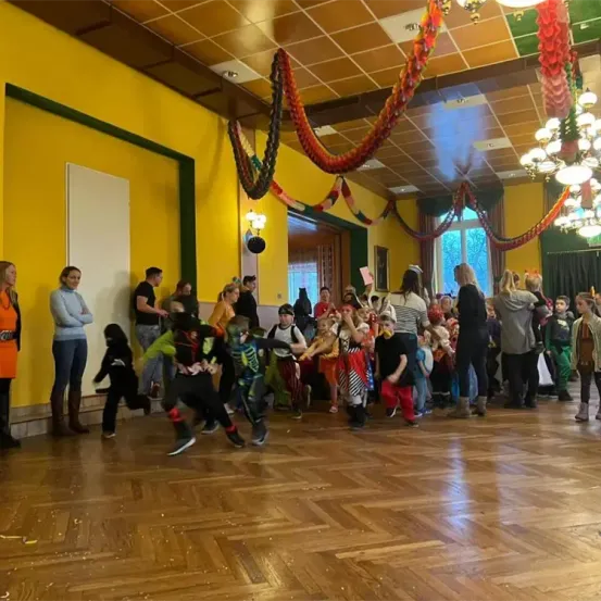 Ein Saal mit Holzboden, Kinder in Kostümen tanzen, während Erwachsene zusehen. Bunte Dekorationen hängen von der Decke.