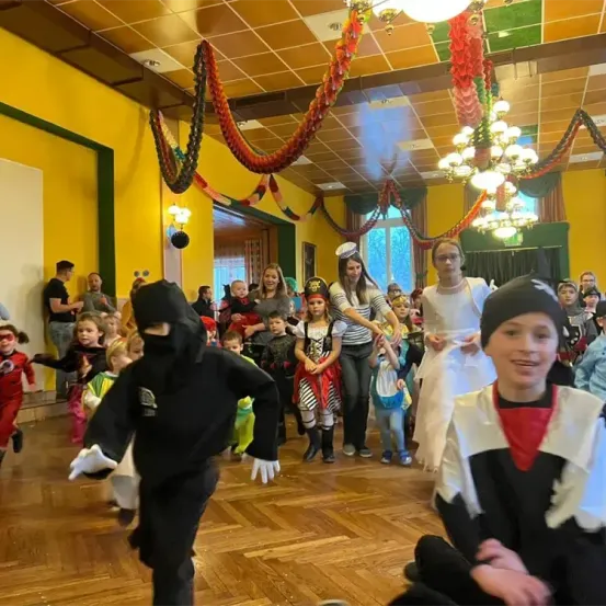 Ein Raum mit Kindern in Kostümen, eines als Ninja verkleidet, feiert mit Erwachsenen. Der Raum hat eine gelbe Wand, Holzboden und Dekorationen.