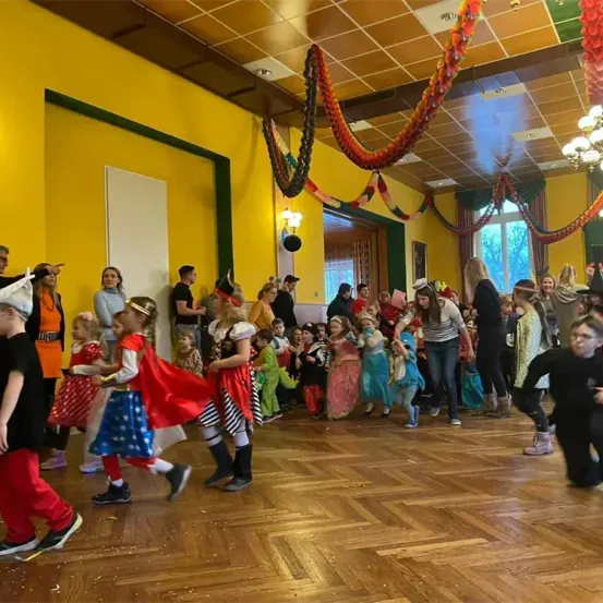 Eine Gruppe von Kindern und Erwachsenen tanzt in Kostümen in einem gelben Raum mit Holzboden und festlicher Dekoration.