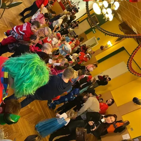 Eine Menschenmenge aus Kindern und Erwachsenen in Kostümen versammelt sich in einem Flur. Viele tragen Rot und Schwarz, einige haben grüne Perücken. Eine Kronleuchter hängt von der Decke.