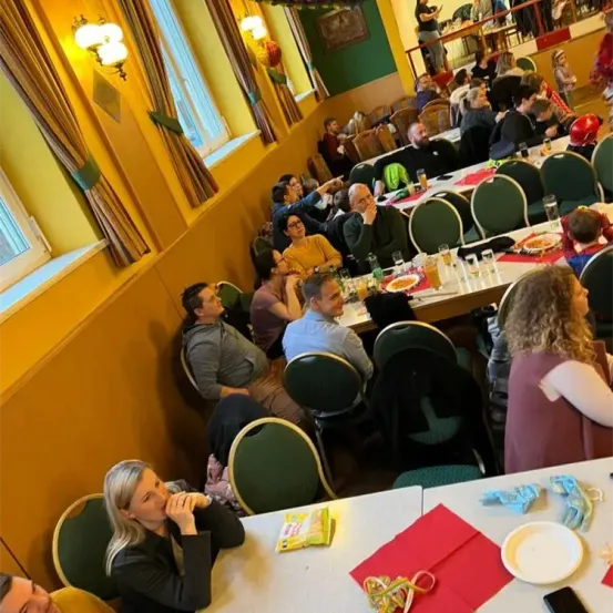 Ein Speisesaal voller Menschen, die an Tischen sitzen, einige essen, andere unterhalten sich. Der Raum ist mit gelben Wänden und Vorhängen dekoriert. Lampen hängen von der Decke, und eine Person steht in der Nähe einer Galerie. Teller, Gläser und Essen stehen auf den Tischen.