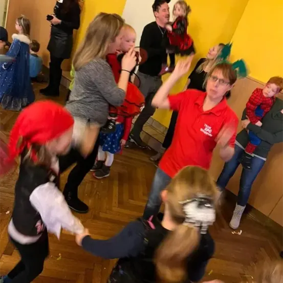 Mehrere Menschen, darunter Kinder, tanzen in einem Raum. Eine Frau in Rot hält ein Kind. Ein Mann mit Brille beobachtet sie. Ein Mädchen mit Piratenhut tanzt mit einem anderen. Eine Frau hält ein Kind auf ihrer Schulter.