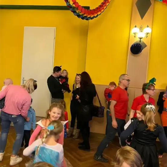 Mehrere Erwachsene und Kinder in einem leuchtend farbigen Raum, einige in Kostümen, tanzen und lächeln. Eine weiße Tür befindet sich links und eine Lampe an der Wand.