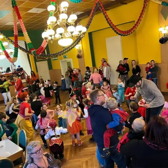 Eine große Gruppe von Menschen in einem Saal, einige in Kostümen, andere in normaler Kleidung. Ein Mann trägt ein Kind, während ein anderer ihn ansieht. Viele Luftballons und eine Kronleuchter schmücken den Raum.