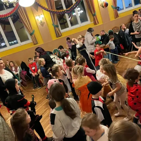 Eine Gruppe von Kindern und Erwachsenen versammelt sich in einem Raum mit Holzboden. Einige Kinder sind in Kostümen gekleidet, und eine Erwachsene hält eine gelbe Schnur. Eine Frau steht und hält ein Baby. Der Raum hat gelbe Wände und Glasfenster.