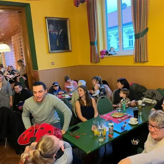 Ein Esszimmer mit gelber Wand, einem gerahmten Bild und einem Fenster mit Vorhängen. Mehrere Personen sitzen an einem grünen Tisch mit Essen und Getränken. Ein Mann in einem Marienkäfer-Kostüm steht neben einer Frau in einem schwarzen Kleid.