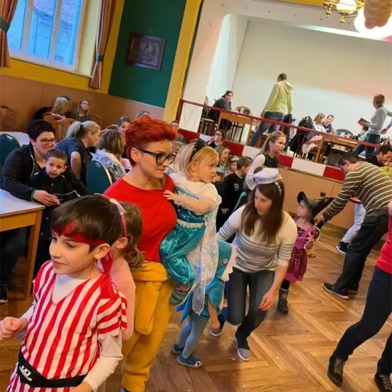 Eine Gruppe von Kindern und Erwachsenen in einem Saal, einige in Kostümen. Eine Frau hält ein Kind in einer Meerjungfrauen-Kostüm. Ein Mann hält die Hand eines Kindes. Leute sitzen an Tischen und auf Bänken im Hintergrund.