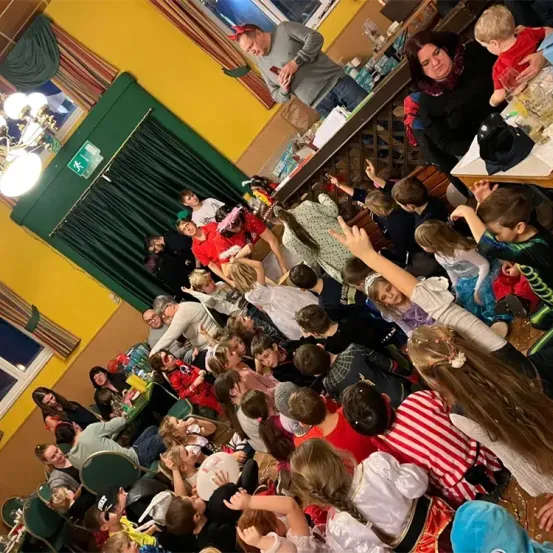 Eine große Gruppe von Kindern und Erwachsenen ist drinnen versammelt, wahrscheinlich bei einer Party oder einem Event. Einige tragen Kostüme und einige sitzen. Eine Laterne und ein Fenster sind im Hintergrund zu sehen.