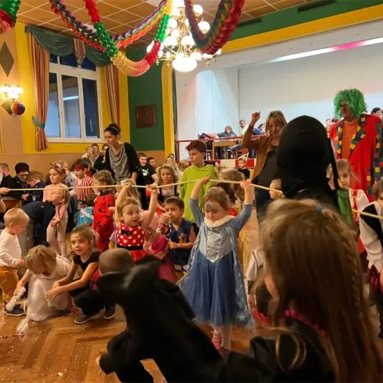 Ein Raum voller Kinder und Erwachsener, viele in Kostümen, halten Seile und beschäftigen sich mit einer festlichen Aktivität. Bunte Dekorationen schmücken den Raum.