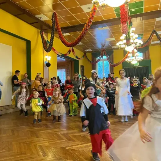 Ein Raum mit Holzboden und gelben Wänden hat eine Party. Eine Kristallleuchte hängt von der Decke, und mehrere Kinder tanzen auf dem Boden. Einige Leute stehen in der Nähe der Wand. Ein paar Kinder tragen Kostüme, und einige tragen Brillen.