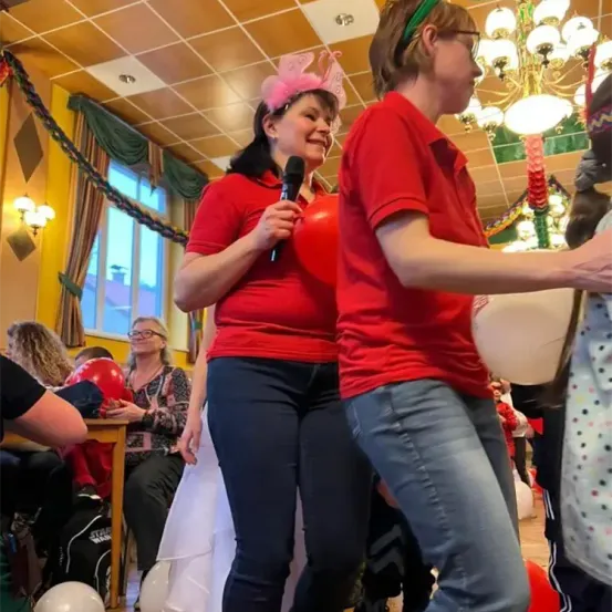 Zwei Personen in roten T-Shirts stehen nebeneinander, eine hält ein Mikrofon. Sie sind in einem Raum mit dekorativen Elementen und Luftballons. Hinter ihnen sitzen und stehen Personen, eine hält einen roten Ballon. Die Umgebung umfasst eine Kronleuchter und ein Fenster.