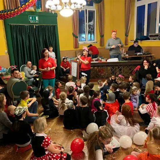 Eine Gruppe von Kindern und Erwachsenen versammelt sich in einem Raum. Die Kinder sind in verschiedenen Kostümen gekleidet. Zwei Frauen stehen vorne, eine hält ein Mikrofon. Der Raum ist mit Luftballons und einem Tisch mit Gegenständen dekoriert.