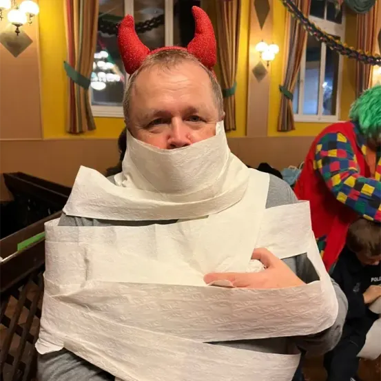 Ein Mann mit einem Teufelshorn-Kopfband und einem mit Toilettenpapier bedeckten Gesicht posiert für ein Foto in einem dekorierten Raum.