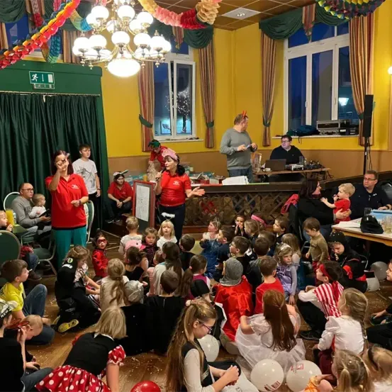 Eine Gruppe von Kindern und Erwachsenen versammelt sich in einem Raum für eine Party. Einige Kinder sitzen auf dem Boden, während andere stehen. Eine Frau in einem roten Shirt hält ein Mikrofon und spricht zu den Kindern. Der Raum ist mit Luftballons und Girlanden dekoriert.