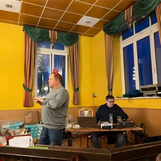 Ein Mann steht vor einem Tisch mit Laptop, trägt eine Stirnband und hält eine Fernbedienung. Ein anderer Mann sitzt an einem Tisch mit diversen Geräten, darunter ein Laptop, und trägt eine Brille. Der Raum hat gelbe Wände und Vorhänge.