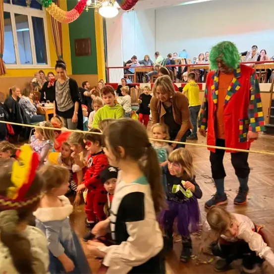 Eine Gruppe von Kindern in Kostümen ist in einem Raum mit Erwachsenen versammelt, wahrscheinlich für eine Party. Einige von ihnen halten Seile, während andere knien. Der Raum hat einen Holzboden und eine weiße Wand. In der Ferne sitzen Menschen auf Stühlen und einige stehen.