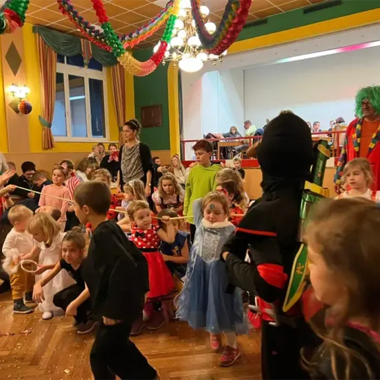 Kinder in Kostümen tanzen in einem Raum mit Holzboden, bunten Dekorationen und Ballons. Ein Kind trägt ein schwarzes Kostüm mit rotem Umhang.