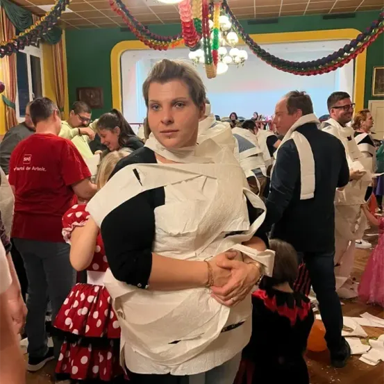 Eine Frau, die in Toilettenpapier gewickelt ist, steht inmitten einer Menschenmenge in einem festlichen Ambiente. Der Raum ist mit bunten Dekorationen und Lichtern geschmückt.