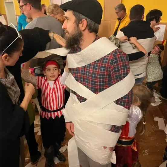 Ein Mann und ein Junge, beide in Toilettenpapier eingewickelt, stehen in einem Raum mit anderen Leuten, von denen einige ebenfalls in Toilettenpapier eingewickelt sind.