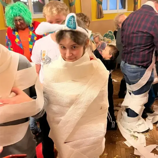 Eine Gruppe von Menschen hat Spaß in einem Raum, wo ein Mädchen in Toilettenpapier eingewickelt ist und Hasenohren auf dem Kopf trägt. Andere Menschen sind ebenfalls in Toilettenpapier eingewickelt.