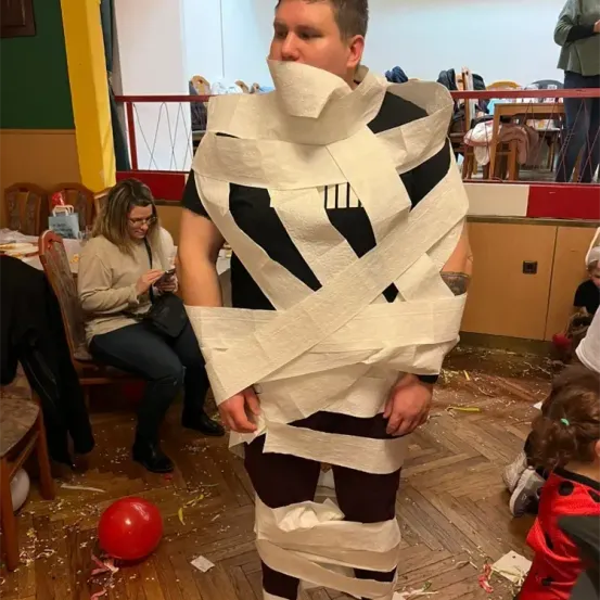 Ein Mann steht in einem Raum und trägt ein Kostüm aus Toilettenpapier. Hinter ihm sitzt eine Frau auf einem Stuhl und ein Kind am Boden. Ein roter Ballon liegt auf dem Boden. Ein Schrank steht rechts, und ein Tisch befindet sich hinter der Frau.