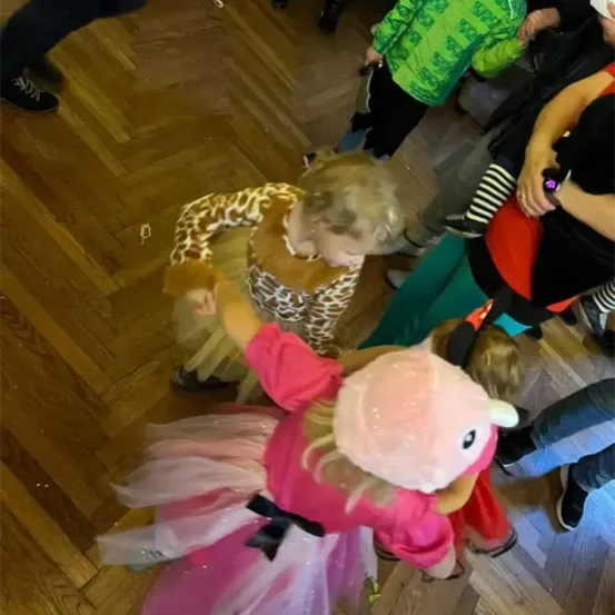 Eine Gruppe von Kindern befindet sich auf einem Holzboden. Ein Mädchen in einem rosa Tutu und Hut blickt nach unten. Ein anderes Kind trägt ein Leoparden-Kostüm. Ein Erwachsener hält ein Kind.