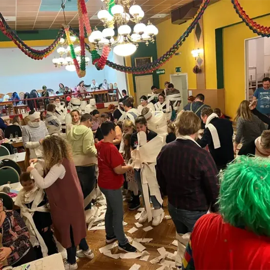 Eine Gruppe von Menschen in einem Raum mit gelben Wänden und Dekorationen, einige in Toilettenpapier gewickelt, lachen und genießen eine festliche Atmosphäre.