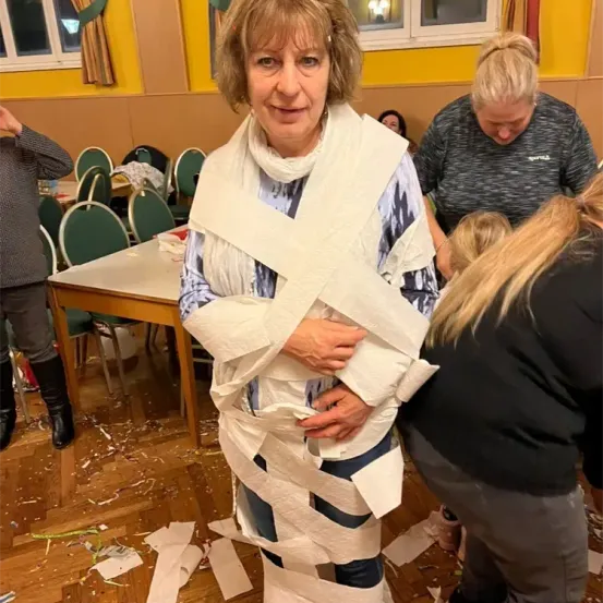 Eine erwachsene Frau, die in Toilettenpapier gewickelt ist, steht in einem Raum mit anderen, die sie anzusehen scheinen. Hinter ihr befindet sich ein Tisch mit Stühlen, und der Boden ist mit Papier übersät.