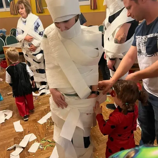 Eine Gruppe von Menschen ist in einem Raum, einige sind in Toilettenpapier eingewickelt, eine Frau in einem roten Kleid hilft einem Kind.