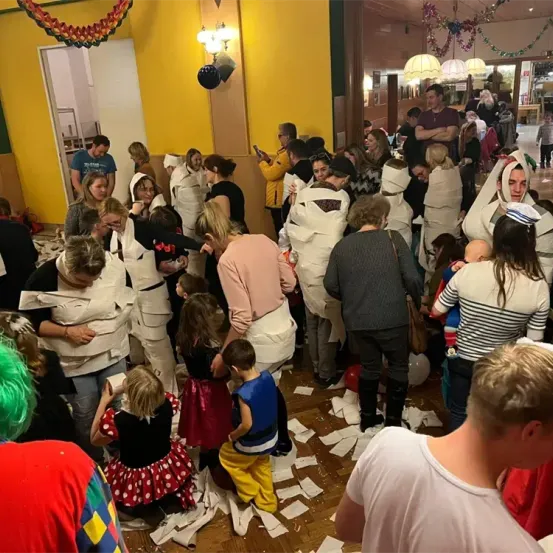 Ein Raum mit gelber Wand und gelben Lichtern. Eine Gruppe von Menschen, einige in Toilettenpapier gekleidet, ist versammelt. Es gibt Kinder und Erwachsene, und einige halten Luftballons. Papiere sind auf dem Boden verstreut.