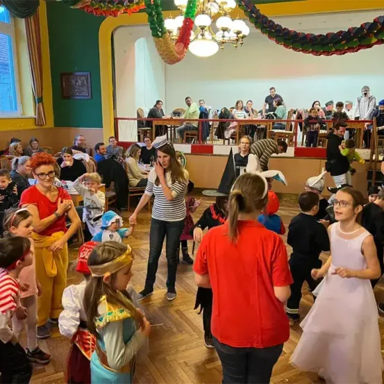 Eine Gruppe von Kindern und Erwachsenen in Kostümen versammelt sich in einem Saal, wahrscheinlich für eine Feier. Eine Frau in der Mitte spricht zu den Kindern. Einige tanzen, während andere zuschauen. Der Saal hat einen Holzboden, eine Kronleuchter und eine rote Brüstung. Einige Leute sitzen und stehen in der Ferne.