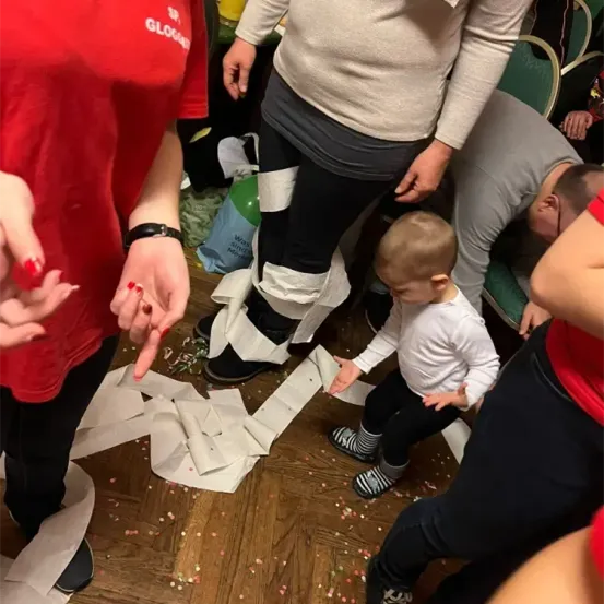 Mehrere Personen befinden sich in einem chaotischen Raum, in dem ein Baby auf dem Boden steht, umgeben von verstreuten Papier und Konfetti. Eine Frau in Rot hält die Hand des Babys, während andere aufräumen.