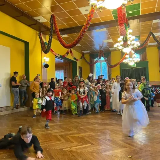 Eine Gruppe von Kindern in Kostümen, einige stehend, einige tanzend, in einem dekorierten Raum. Ein Mädchen in einem weißen Kleid geht nach vorne.