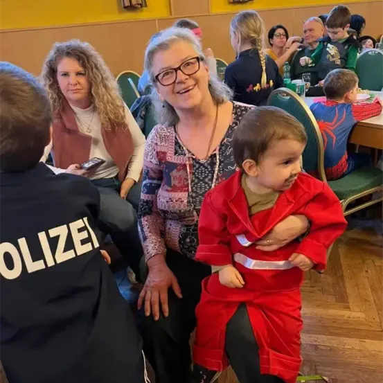Eine Frau sitzt mit einem kleinen Kind in einem roten Overall. Sie sind an einem Tisch mit einem Polizisten. Andere Leute sitzen hinter ihnen.