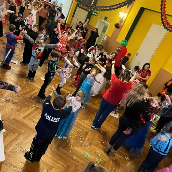 Eine Gruppe von Kindern in verschiedenen Kostümen tanzt in einem Raum. Einige halten Schwerter und Zauberstäbe. Der Raum hat einen Holzboden und gelbe Wände.